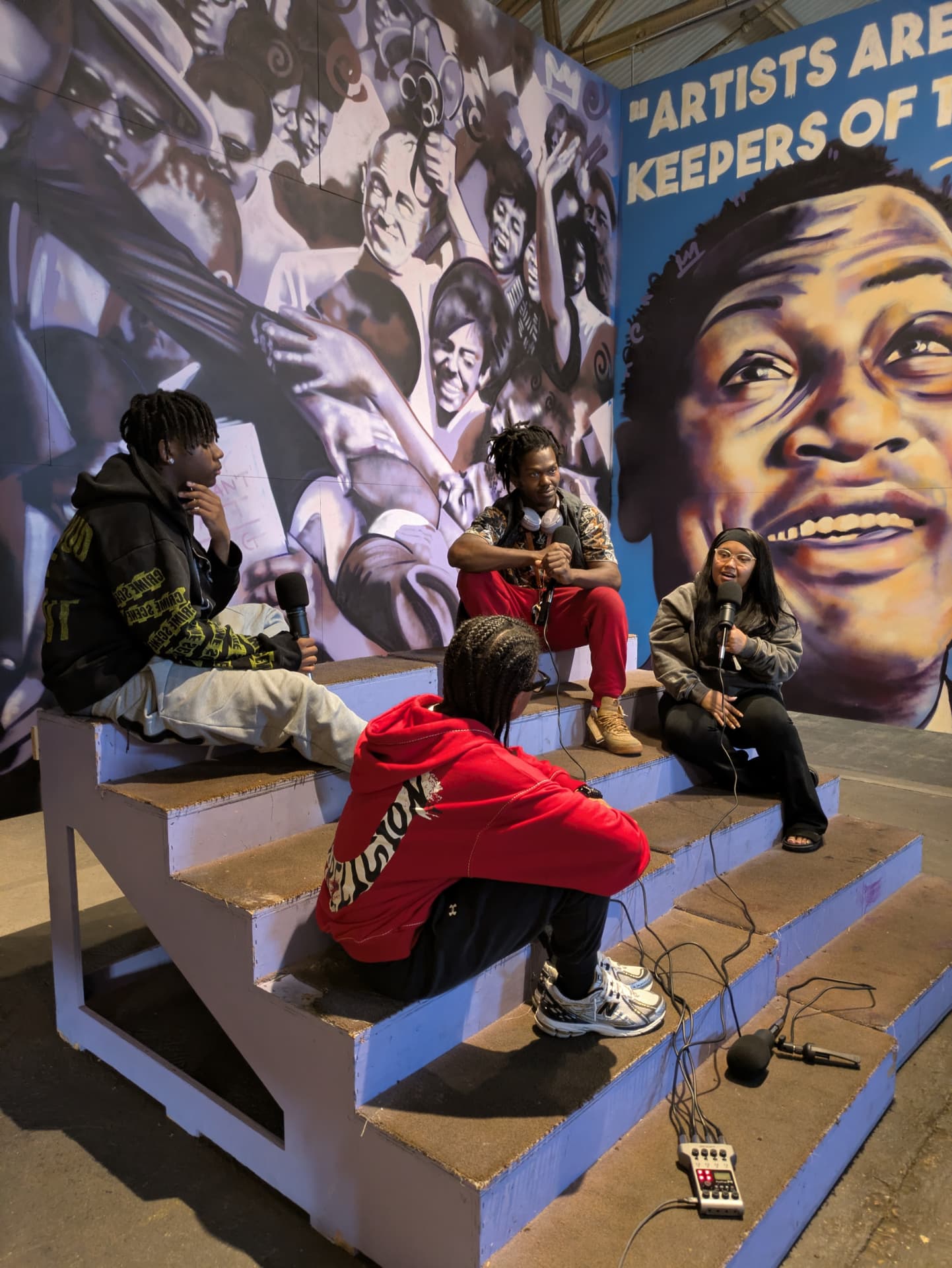 Four young DJs recording with handheld mics on steps in front of a large community mural