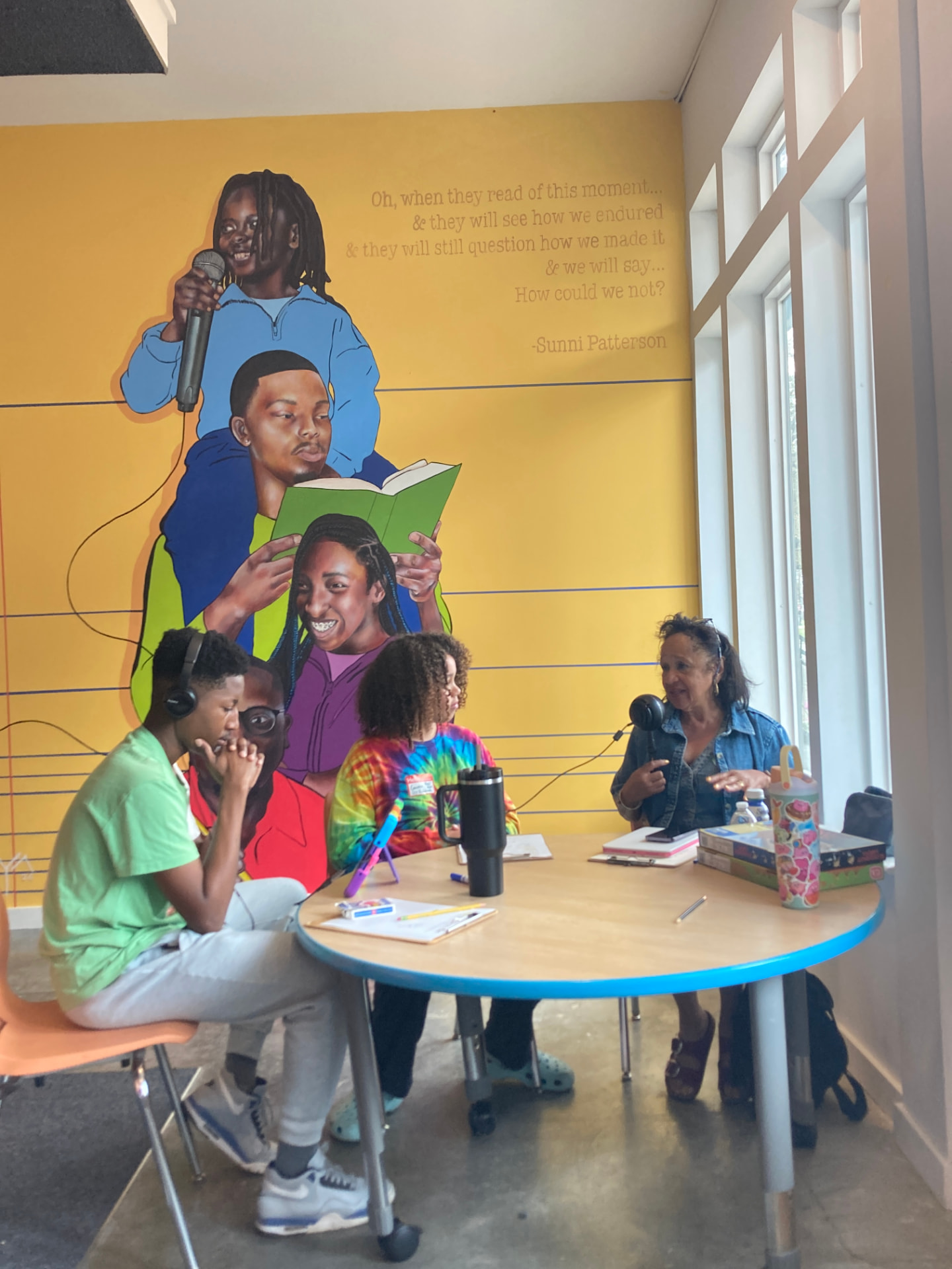 Three people gathered around a blue-rimmed table recording a podcast segment in front of a yellow mural wall